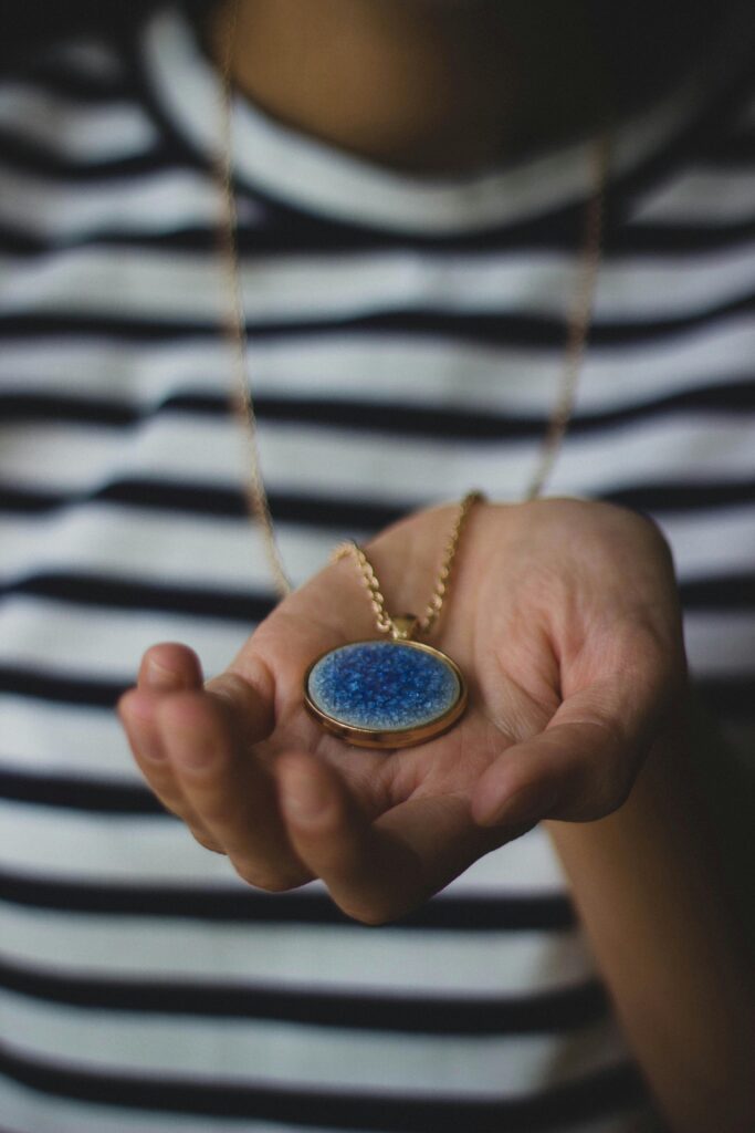 Close-up of a hand holding a blue pendant necklace against a striped shirt.